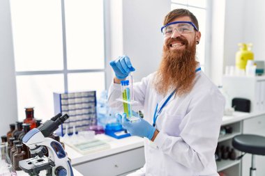 Laboratuvarda test tüpleri tutan bilim adamı üniforması giyen genç kızıl saçlı adam.