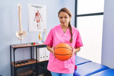 Fizyoterapi kliniğinde çalışan genç İspanyol kadın basketbol topunu kuşkucu ve gergin tutuyor, sorun yüzünden surat asıyor. negatif kişi. 