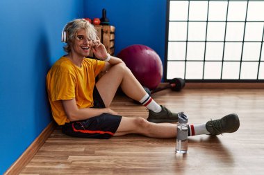 Genç sarışın adam spor merkezinde yerde oturmuş müzik dinliyor.