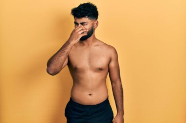 Arab man with beard wearing swimwear shirtless smelling something stinky and disgusting, intolerable smell, holding breath with fingers on nose. bad smell 