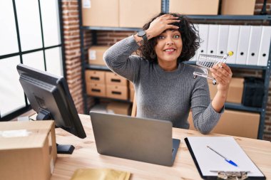 Hispanic doctor woman with curly hair standing by manikin at small business stressed and frustrated with hand on head, surprised and angry face 