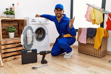 Young indian technician working on washing machine with a big smile on face, pointing with hand finger to the side looking at the camera. 