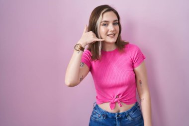 Blonde caucasian woman standing over pink background smiling doing phone gesture with hand and fingers like talking on the telephone. communicating concepts. 