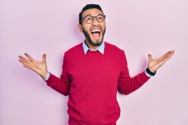 Hispanic man with beard wearing business shirt and glasses crazy and mad shouting and yelling with aggressive expression and arms raised. frustration concept. 