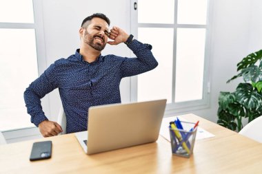 Young hispanic man with beard working at the office with laptop stretching back, tired and relaxed, sleepy and yawning for early morning 