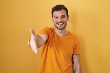 Sarı arka planda duran Latin asıllı genç adam dostça bir selamlama ve hoş geldin hediyesi olarak el sıkışmayı sunuyor. başarılı iş. 