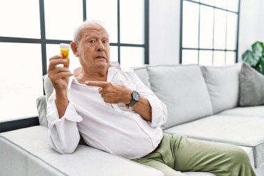 Senior man holding pills pointing aside worried and nervous with forefinger, concerned and surprised expression 