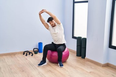 Young hispanic man sitting on fit ball stretching at sport center