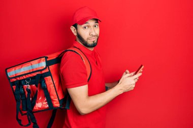 Hispanic man with beard wearing delivery package backpack using smartphone clueless and confused expression. doubt concept. 