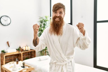 Redhead man with long beard wearing bathrobe at wellness spa screaming proud, celebrating victory and success very excited with raised arms 