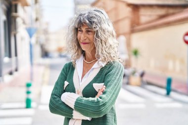 Orta yaşlı kadın gülümsüyor. Kollarını kavuşturup caddede duruyor.