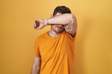 Sarı arka planda dikilen İspanyol genç adam kollarıyla gözlerini kapatıyor, ciddi ve üzgün görünüyor. Görme, saklama ve reddetme konsepti 