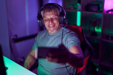 Beyaz saçlı İspanyol bir adam akıllı telefonuyla video oyunu oynuyor. Olumlu ve mutlu görünüyor. Kendine güvenen bir gülümsemeyle dişlerini gösteriyor. 