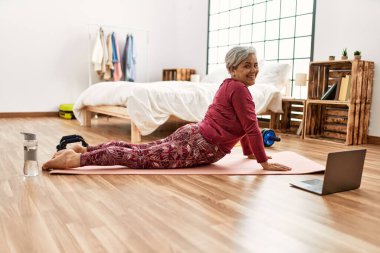 Orta yaşlı, kır saçlı bir kadın. Yatak odasında yerde uzanan internetten esneme dersleri alıyor..