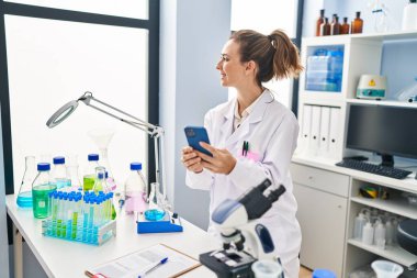 Laboratuvarda akıllı telefon kullanan bilim adamı üniforması giyen genç bir kadın.