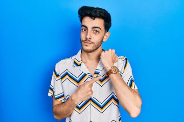 Gündelik kıyafetler giyen genç İspanyol adam saati, sabırsızlığı ve rahat bir ifadeyle kameraya bakmayı işaret ediyor. 