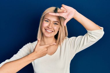 Beautiful blonde woman wearing casual sweater smiling making frame with hands and fingers with happy face. creativity and photography concept. 