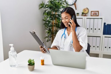 Doktor üniforması giyen genç Latin kadın klinikte akıllı telefondan konuşuyor.