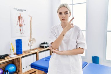 Young caucasian woman working at pain recovery clinic pointing with hand finger to the side showing advertisement, serious and calm face 
