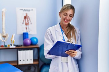 Fizyoterapi kliniğinde kontrol listesine yazılı fizyoterpist üniforması giyen genç sarışın kadın.