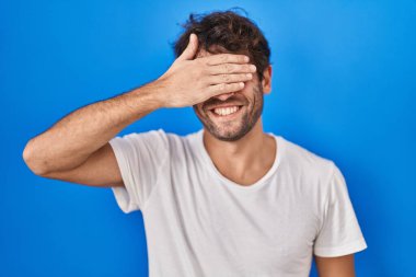 Latin kökenli genç adam mavi arka planda duruyor. Gülümsüyor ve gülümsüyor. Gözleri şaşkınlıkla örtüyor. kör kavram. 