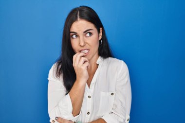 Mavi arka planda dikilen genç İspanyol kadın gergin ve gergin görünüyor. Elleri ağızdan tırnak yiyor. anksiyete. 
