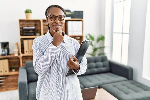 African woman working at psychology clinic looking confident at the camera smiling with crossed arms and hand raised on chin. thinking positive.