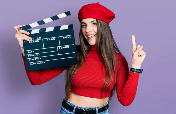 Young brunette teenager holding video film clapboard smiling with an idea or question pointing finger with happy face, number one