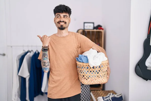 Young hispanic man with beard holding laundry basket at bedroom pointing thumb up to the side smiling happy with open mouth