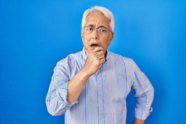 Hispanic senior man wearing glasses looking fascinated with disbelief, surprise and amazed expression with hands on chin