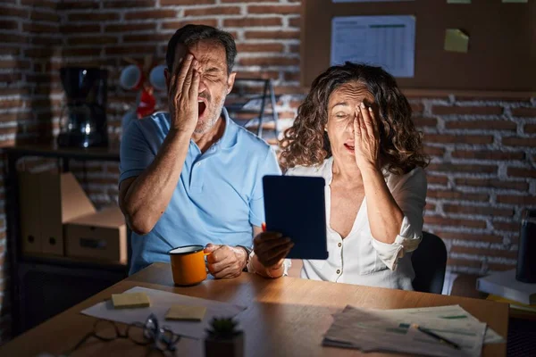 Middle age hispanic couple using touchpad sitting on the table at night yawning tired covering half face, eye and mouth with hand. face hurts in pain.