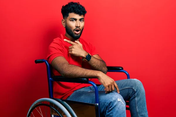 Arab man with beard sitting on wheelchair surprised pointing with finger to the side, open mouth amazed expression.