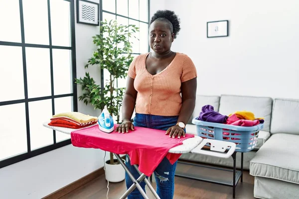 Young african woman ironing clothes at home puffing cheeks with funny face. mouth inflated with air, crazy expression.