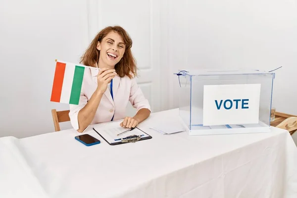 Beautiful caucasian woman at political campaign election holding hungary flag looking positive and happy standing and smiling with a confident smile showing teeth