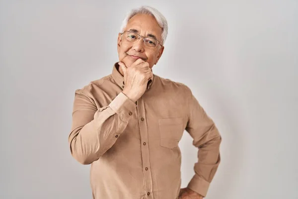 Hispanic senior man wearing glasses looking confident at the camera with smile with crossed arms and hand raised on chin. thinking positive.
