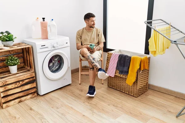 Young Hispanic Man Waiting Washing Machine Using Smartphone Laundry Room — стоковое фото