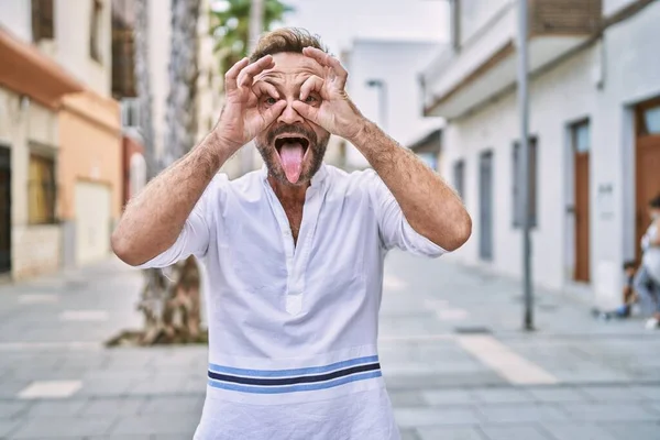 Middle age man outdoor at the city doing ok gesture like binoculars sticking tongue out, eyes looking through fingers. crazy expression.