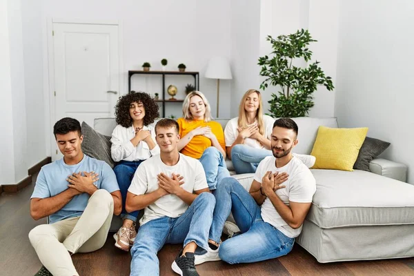 Group of people sitting on the sofa and floor at home smiling with hands on chest with closed eyes and grateful gesture on face. health concept.