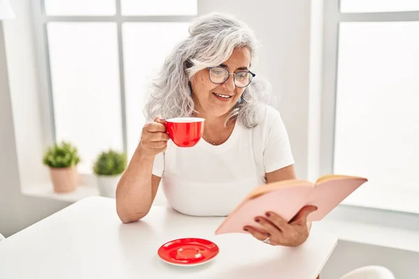 Middle Age Woman Drinking Coffee Reading Book Sitting Table Home — Stok fotoğraf