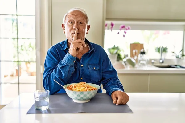 Senior man with grey hair eating pasta spaghetti at home asking to be quiet with finger on lips. silence and secret concept.