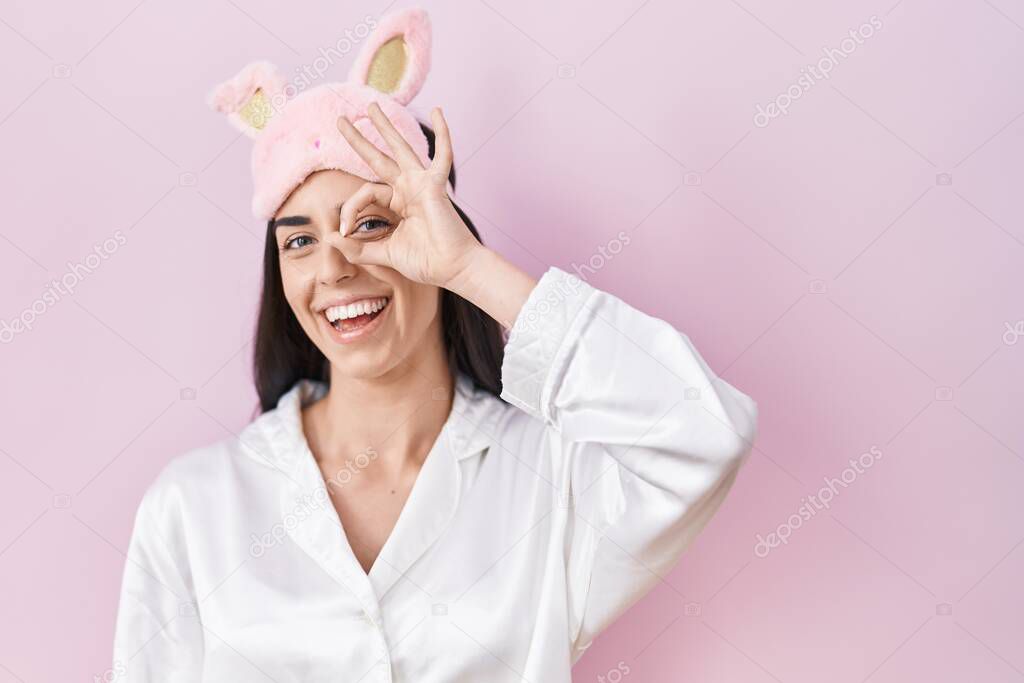Young brunette woman wearing sleep mask and pajama smiling happy doing ok sign with hand on eye looking through fingers 