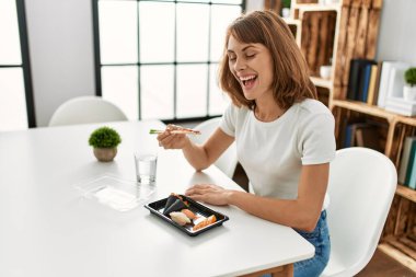 Genç beyaz kadın suşi yiyor. Evdeki masada oturuyor.