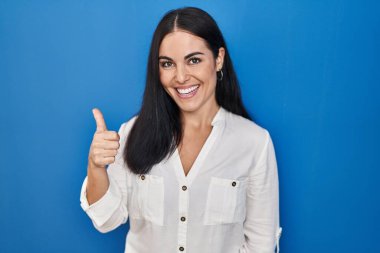 Mavi arka planda dikilen genç İspanyol kadın elleriyle mutlu bir jest yapıyor. Kameraya bakıp başarıyı gösteren ifade. 
