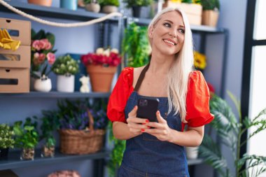 Young blonde woman florist smiling confident using smartphone at florist store
