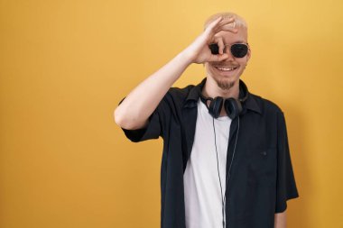 Young caucasian man wearing sunglasses standing over yellow background doing ok gesture with hand smiling, eye looking through fingers with happy face. 