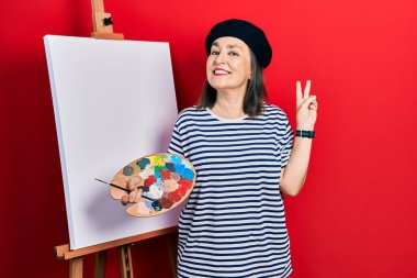 Middle age hispanic woman standing drawing with palette by painter easel stand smiling looking to the camera showing fingers doing victory sign. number two. 