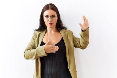 Young brunette woman standing over isolated background swearing with hand on chest and open palm, making a loyalty promise oath 