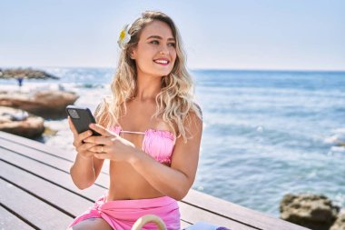 Kumsalda bankta oturan akıllı telefon kullanan genç sarışın kız..