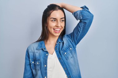 Hispanic woman standing over blue background smiling confident touching hair with hand up gesture, posing attractive and fashionable 