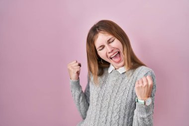 Güzel bir kadın pembe arka planda mutlu ve heyecanlı bir şekilde kollarını kaldırıp, gülümseyerek ve başarı için çığlık atarak zafer jesti yapıyor. kutlama konsepti. 